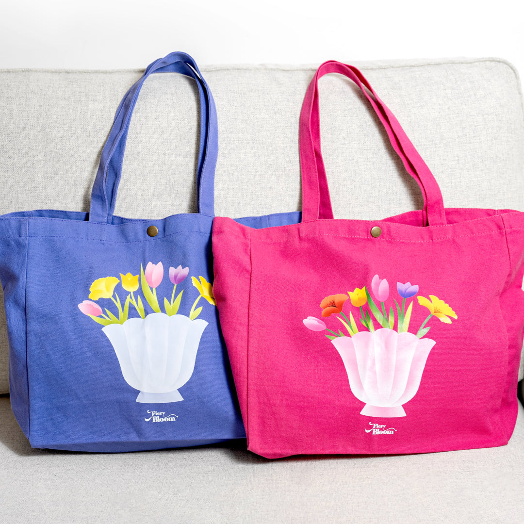 Two canvas tote bags in pink and purple side-by-side, both featuring a shell-shaped vase with tulip designs.
