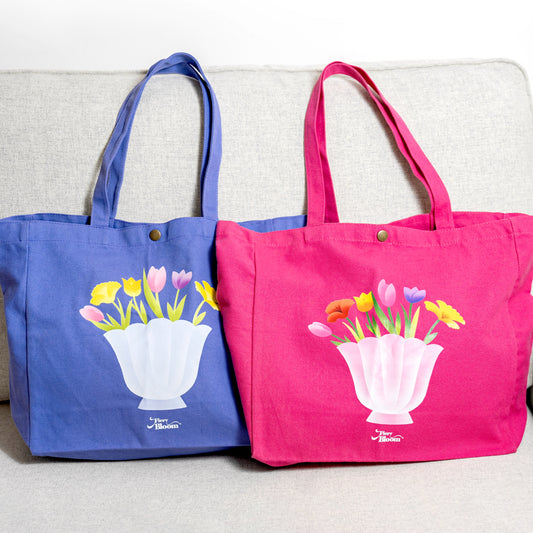 Two canvas tote bags in pink and purple side-by-side, both featuring a shell-shaped vase with tulip designs.