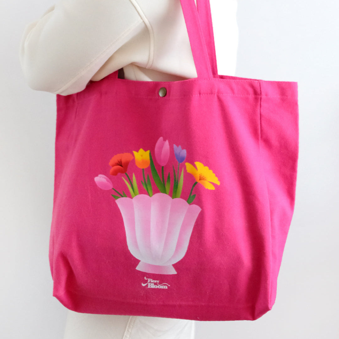 Pink canvas tote bag featuring an artistic illustration of a shell-shaped vase with vibrant pink and yellow tulips.